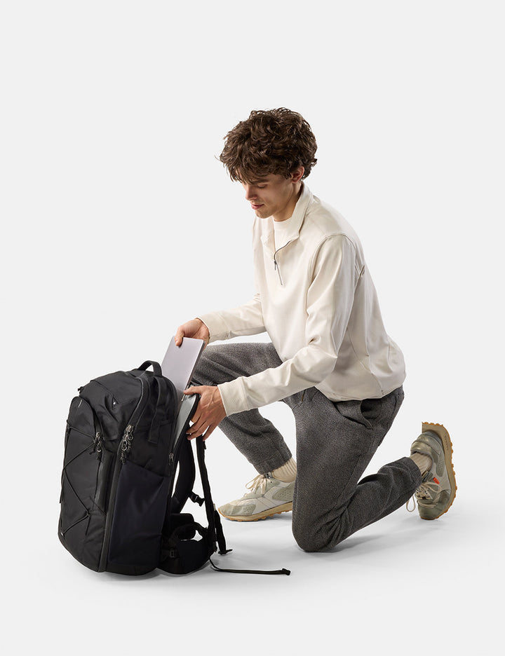 Matador 35L Global Travel Pack in Black - man sitting on the floor with a black backpack and laptop on a white background