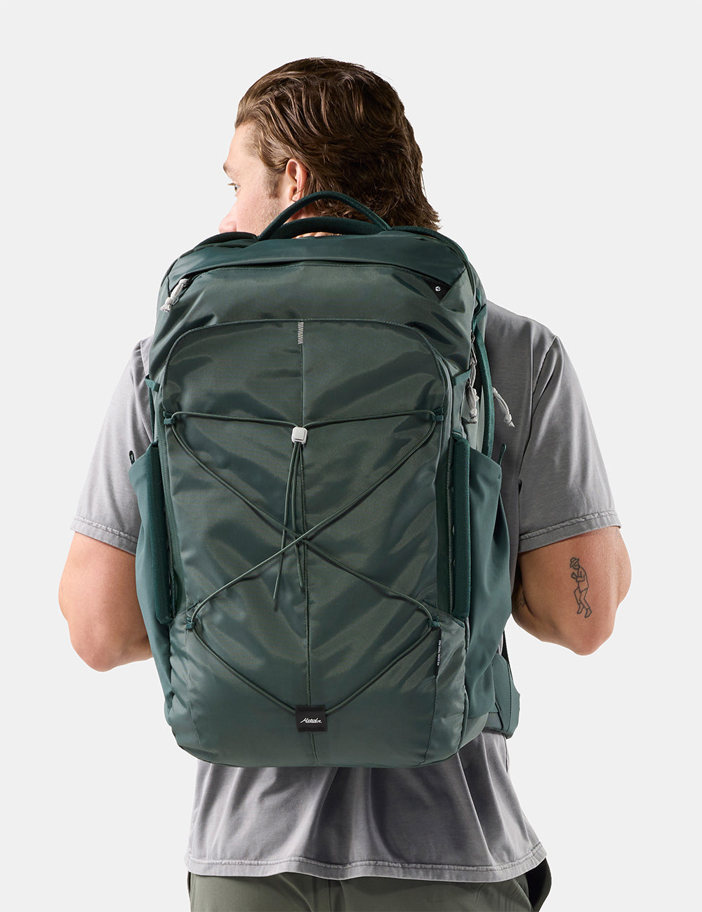 Matador 45L Global Travel Pack in Juniper - a man viewed from behind wearing a dark green technical backpack with multiple zippered compartments and compression straps, set against a plain light gray studio background