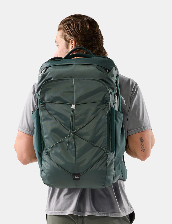 Matador 45L Global Travel Pack in Juniper - a man viewed from behind wearing a dark green technical backpack with multiple zippered compartments and compression straps, set against a plain light gray studio background