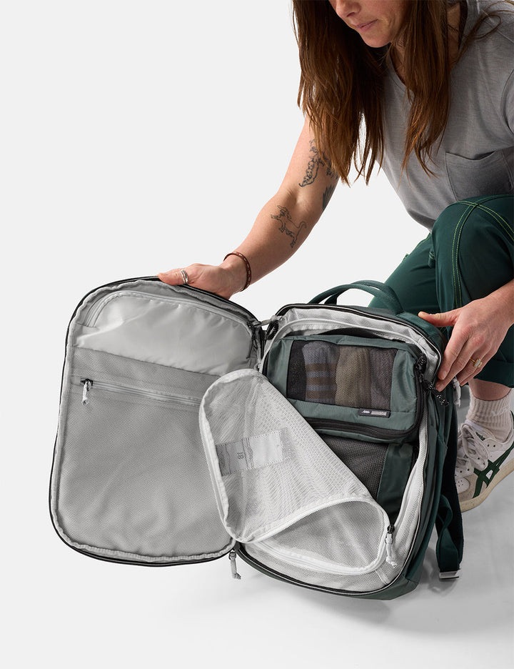 Matador 18L Personal Item Travel Pack in Juniper - person opening a green travel backpack with a clear front pocket on a white background