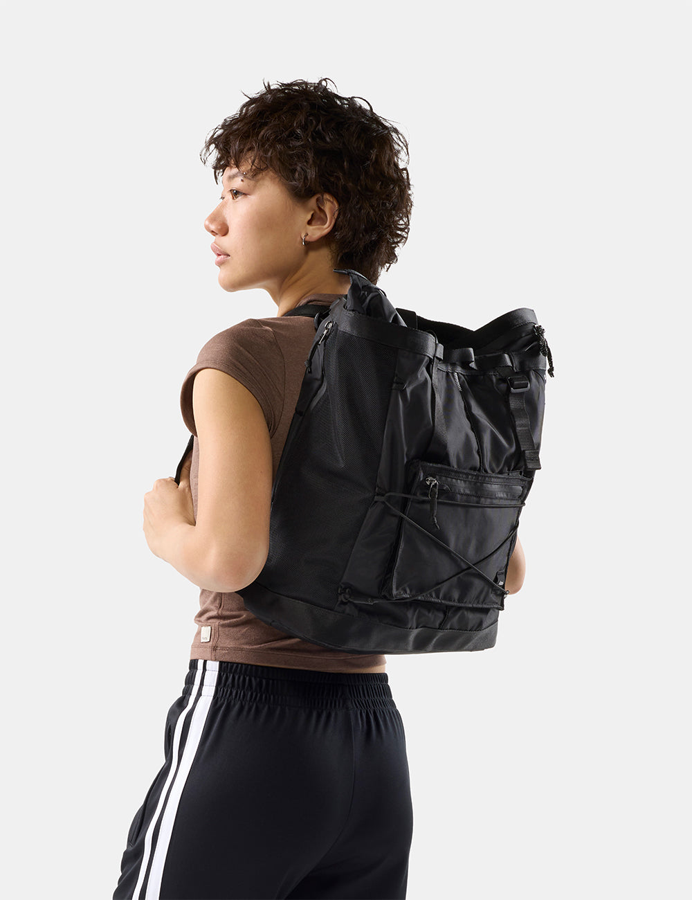 Matador 24L Tote Pack in Black - a person wearing a black backpack with multiple zippered pockets and compression straps. They are wearing a brown sleeveless top and black athletic pants with white stripes, photographed against a clean light gray studio background